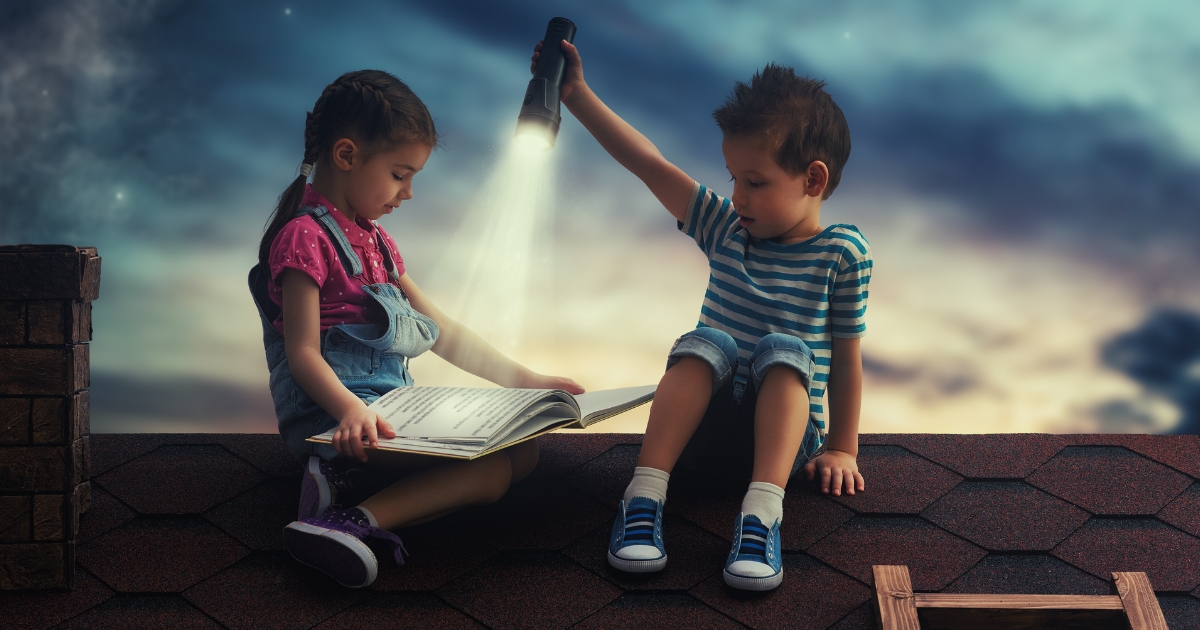 子ども2人が本を屋根に上がり、家中電灯で本を照らし、学ぶ様子の画像。